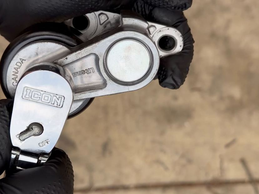 Gloved hands holding a ratchet on a belt tensioner over concrete